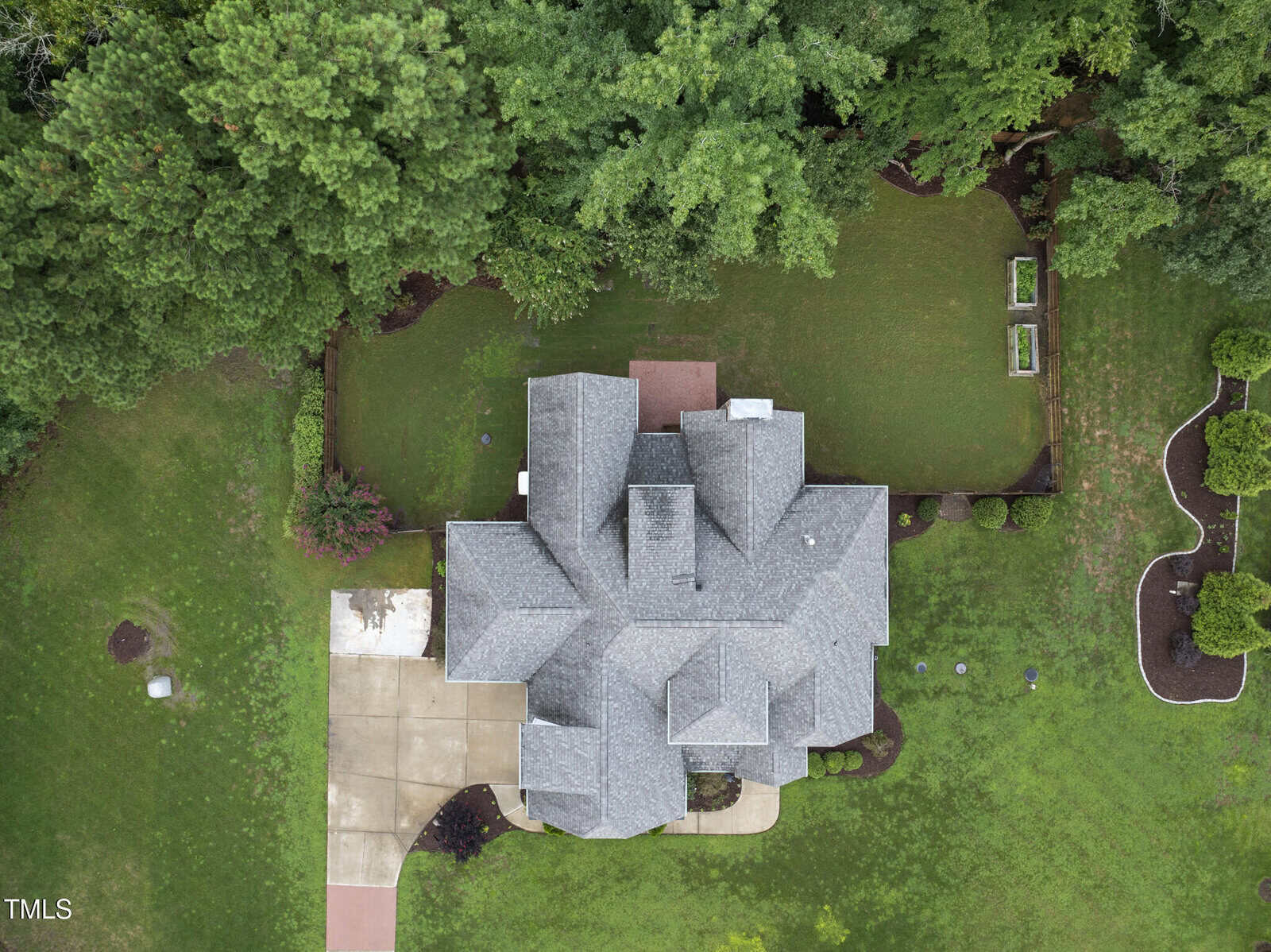 Aerial view of a house with a gray roof, surrounded by green lawn and trees. The driveway is visible on the left, and garden beds and landscaping are on the right. The backyard is partially fenced with neatly trimmed grass.
