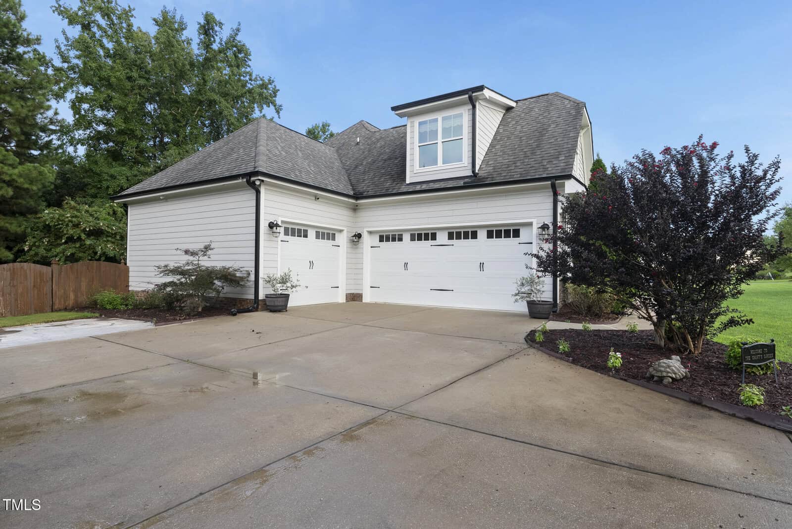 A spacious, modern three-car garage attached to a white house with gray shingles, surrounded by landscaped greenery, shrubs, and a clean driveway on a sunny day.