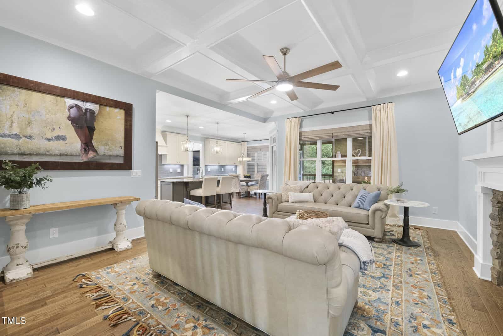 Bright living room with light blue walls, coffered ceiling, beige tufted sofa, patterned rug, and wall-mounted TV. Large window with curtains, open layout leading to kitchen, and decorative consoles and artwork on the walls.