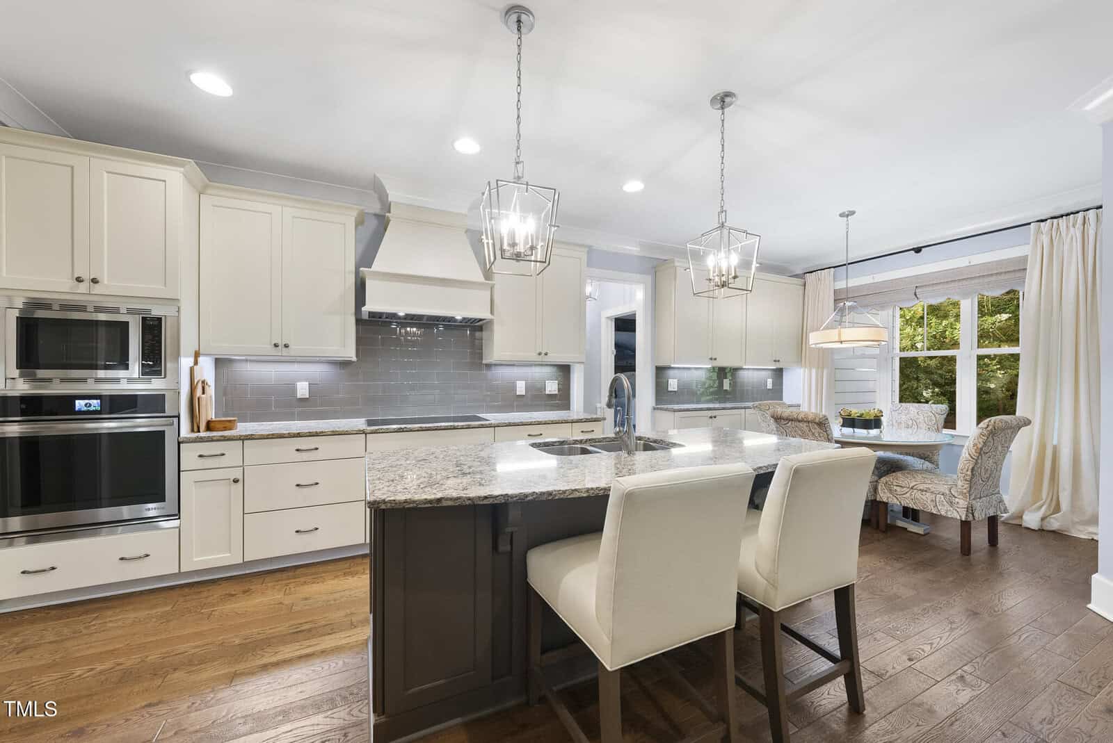 A modern kitchen with white cabinets, stainless steel appliances, a gray tile backsplash, granite island with sink, two cushioned barstools, pendant lights, and a dining nook by a large window with beige curtains.