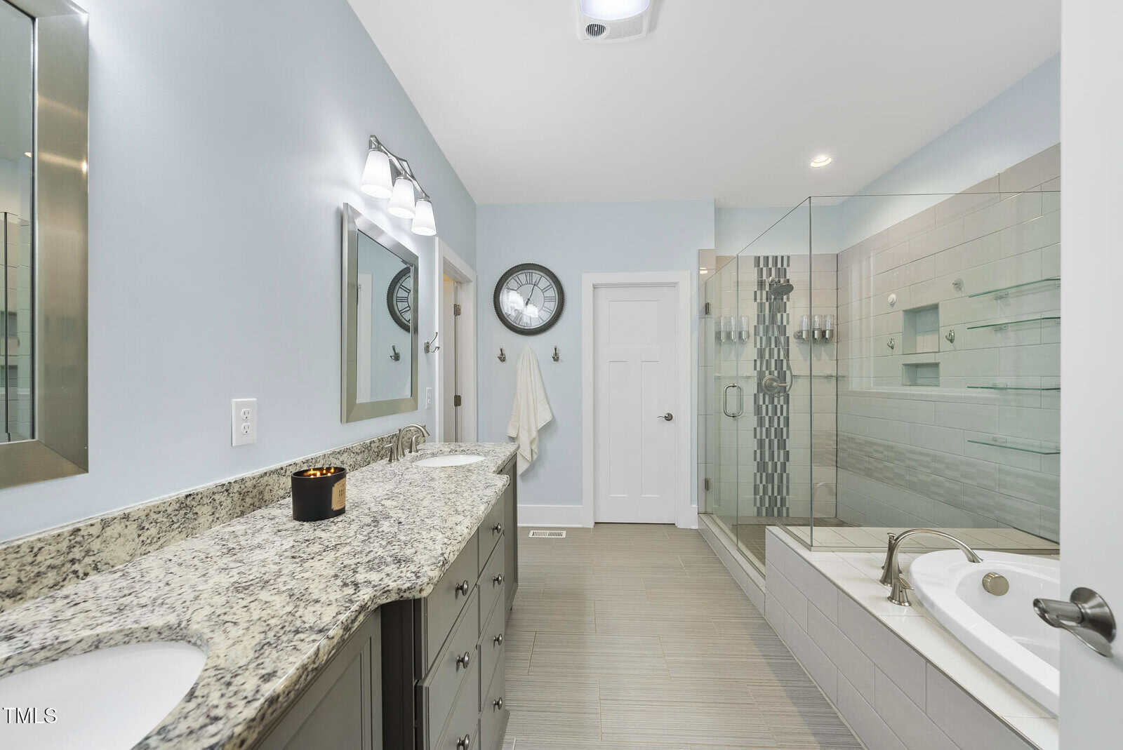 Modern bathroom with double granite sink vanity, wall-mounted mirrors, glass-enclosed shower with tile accents, bathtub with faucet, light blue walls, and a wall clock above a towel. The floor is beige tile.