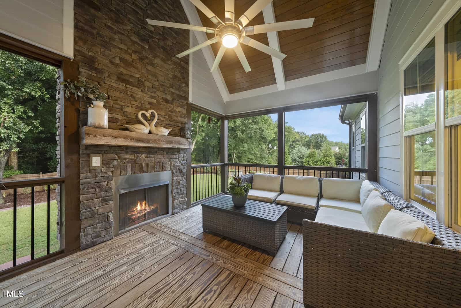 Screened-in porch with a stone fireplace, wooden ceiling, and deck floor, featuring a ceiling fan, wicker sectional sofa with cushions, a coffee table, and large windows overlooking a green yard.
