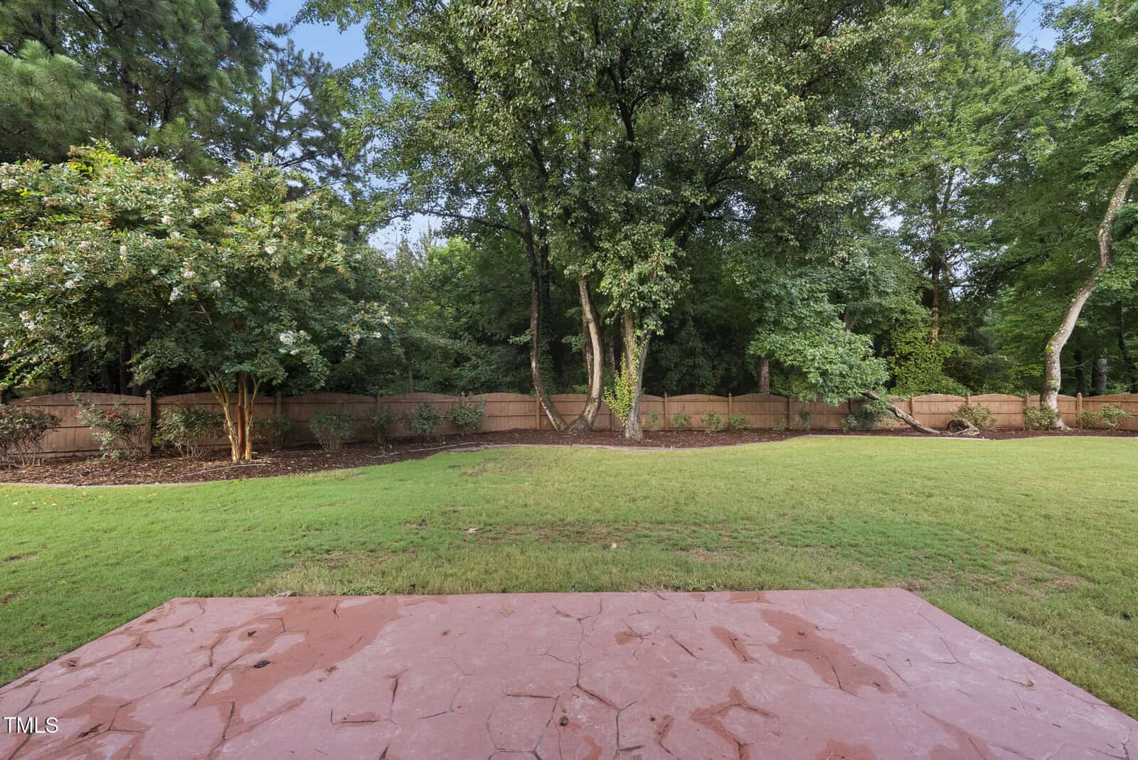 View from a stone patio looking out onto a large green backyard with a wooden fence and various mature trees and shrubs bordering the lawn. The sky is not visible.