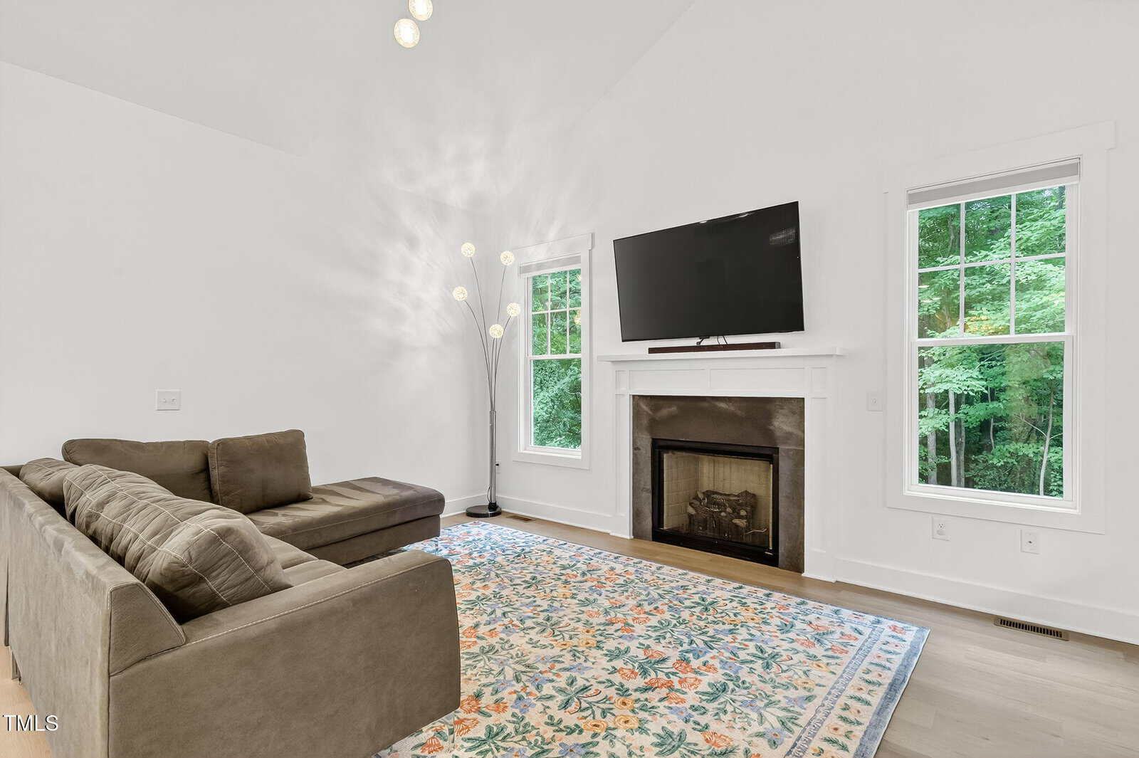 A living room with a sectional sofa, patterned rug, floor lamp, fireplace with a TV mounted above, two windows, and white walls. The windows show greenery outside.