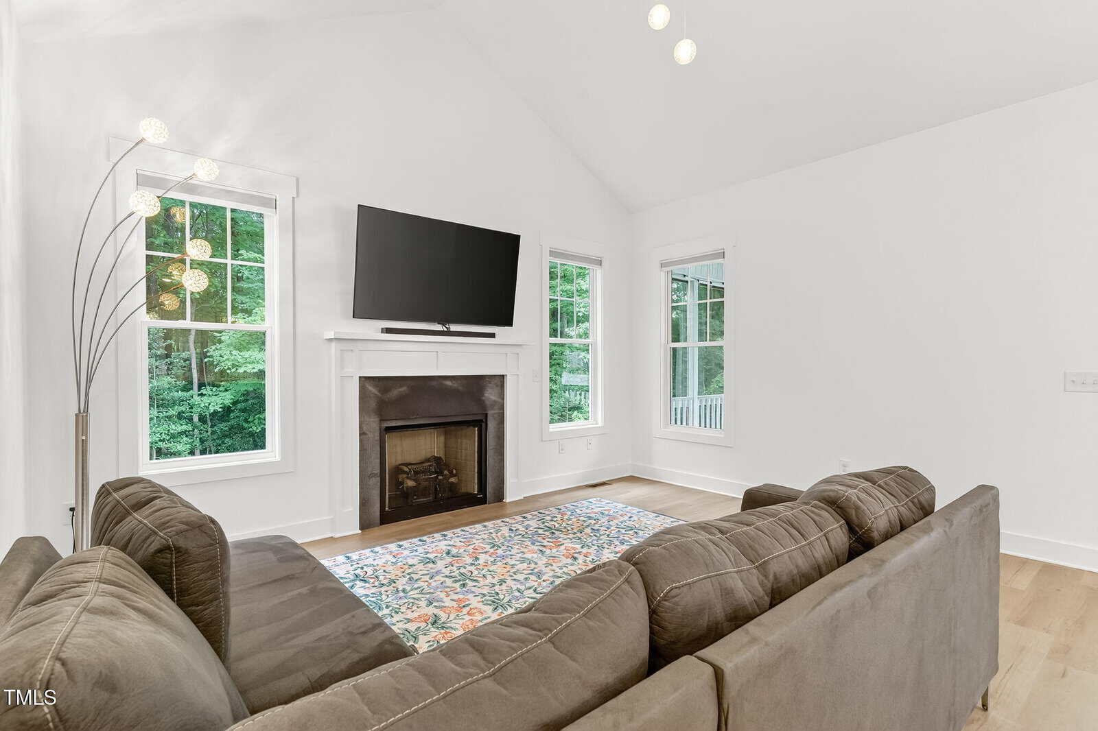 A living room with a brown sectional sofa, a fireplace with a TV mounted above it, two windows with views of greenery, a patterned rug, and a modern floor lamp. The walls and ceiling are white.