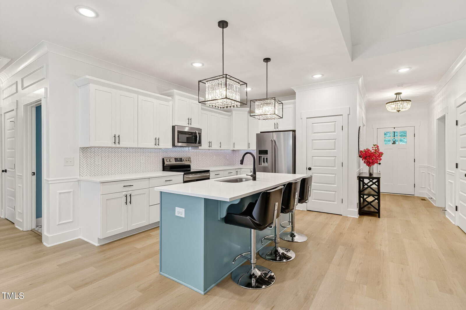 Modern white kitchen with stainless steel appliances, a large island with a blue base, black bar stools, geometric pendant lights, and light wood flooring. A small table with red flowers is near the entryway.