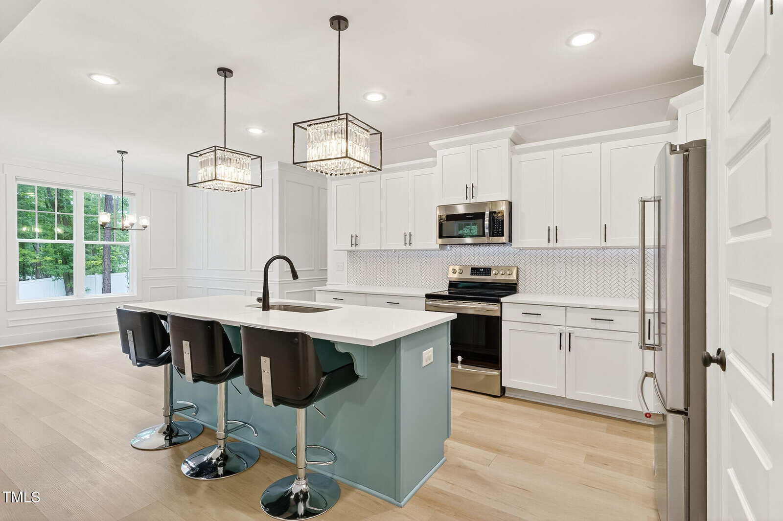 Modern kitchen with white cabinets, stainless steel appliances, a large island with a sink, three black barstools, pendant lights, light wood floors, and a dining area with large windows in the background.