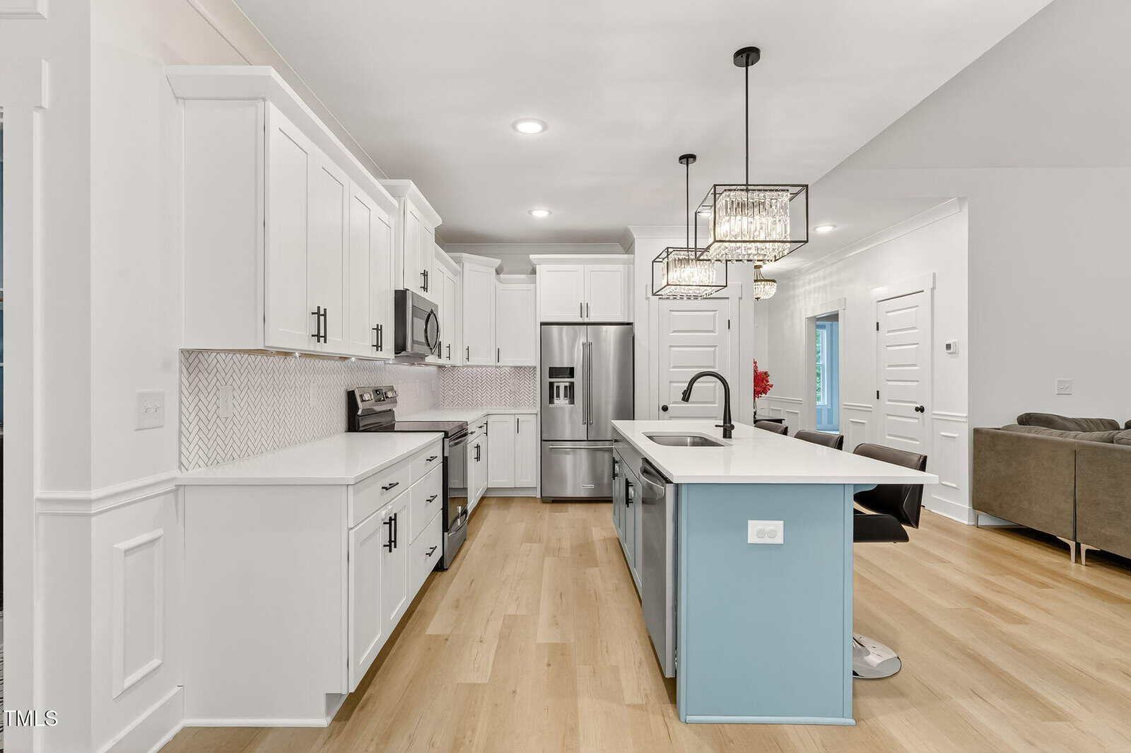 Modern kitchen with white cabinets, stainless steel appliances, a large island with a light blue base and white countertop, pendant lights above, light wood flooring, and an open layout leading to a living area.