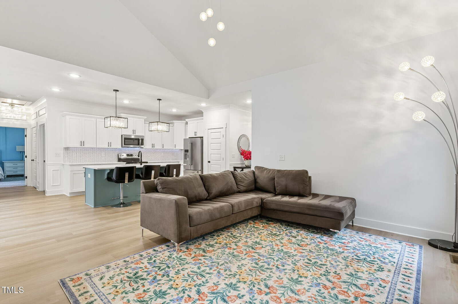 Modern living room with a gray sectional sofa on a colorful floral rug, open to a kitchen with white cabinets, island seating, and stainless steel appliances. A floor lamp with multiple lights stands in the corner.