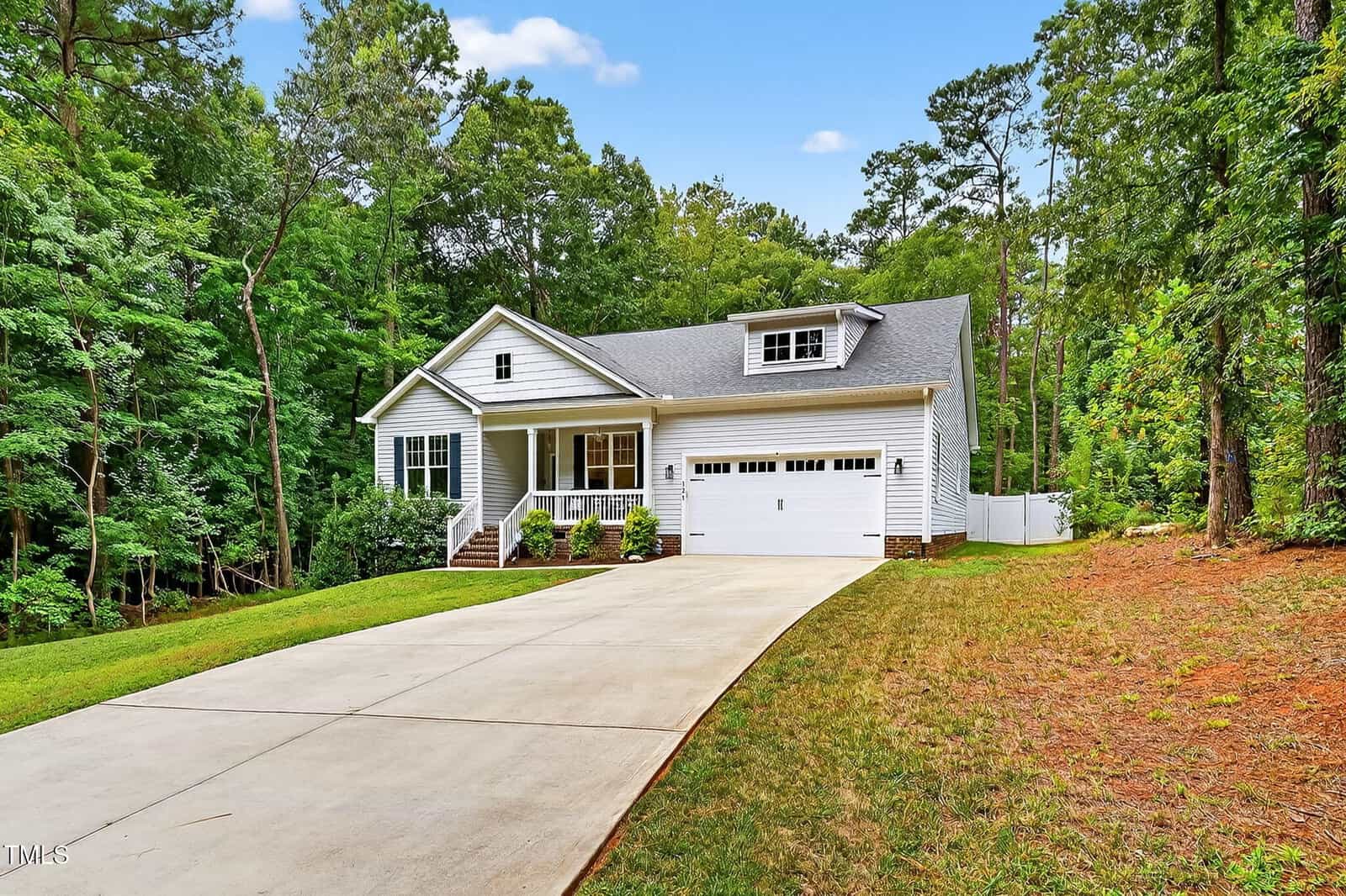 A white single-story house with a front porch, attached two-car garage, and a long driveway, surrounded by tall green trees and a grassy yard under a blue sky.