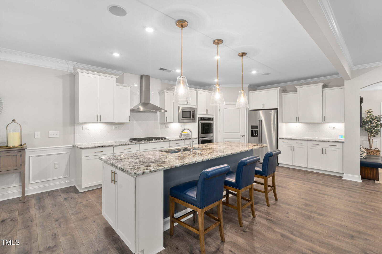 Modern kitchen with white cabinets, granite countertops, stainless steel appliances, a large island with three blue chairs, pendant lighting, and wood flooring. The space is bright with a clean, open layout.