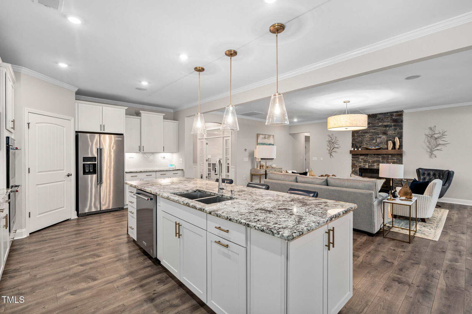 Modern open-concept kitchen and living area with white cabinets, stainless steel appliances, granite island, wood floors, and pendant lights. The living room features a stone fireplace and neutral furnishings.