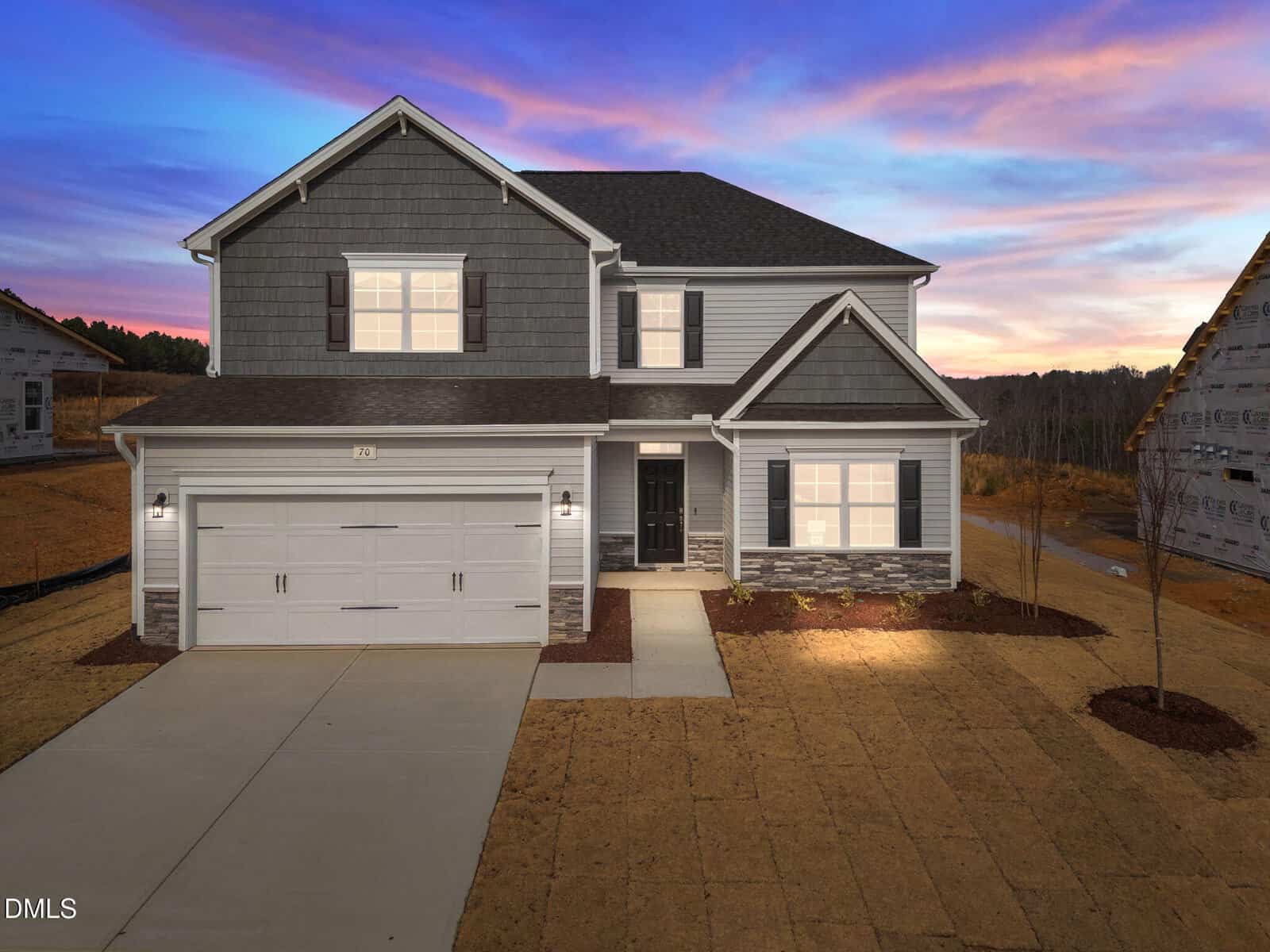 A two-story suburban house with gray siding, white trim, a double garage, and a black front door. The yard is unlandscaped, and the sky is colorful at sunset with pink and purple tones.