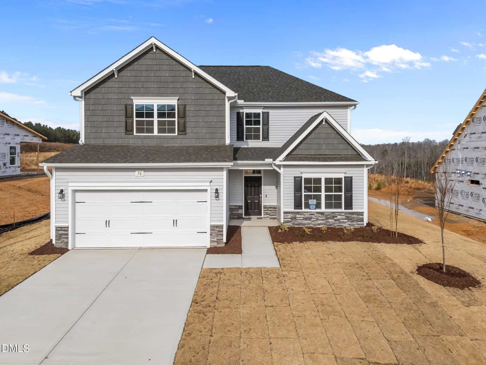 Newly constructed two-story house with gray siding, double garage, paved driveway, and a small front porch. The yard has freshly laid sod and minimal landscaping, with similar homes visible nearby under development.