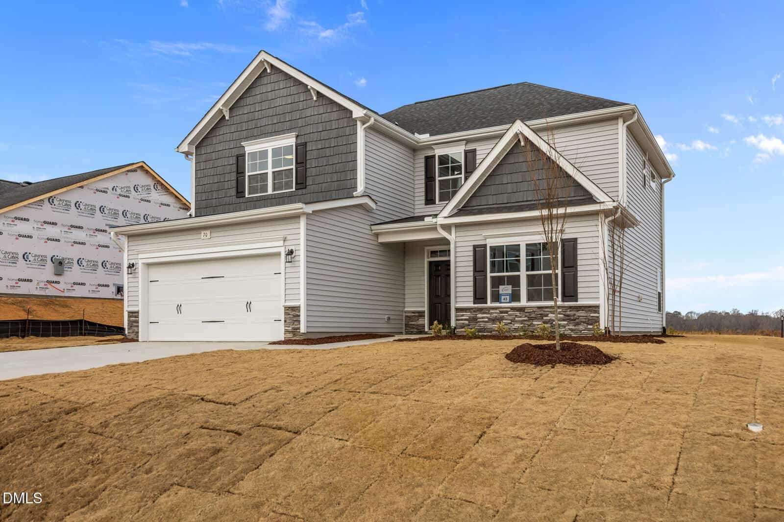 Two-story modern suburban house with gray siding, dark shingles, and white trim. Attached two-car garage and a newly landscaped yard with fresh sod. Neighboring house under construction is visible on the left. Blue sky overhead.