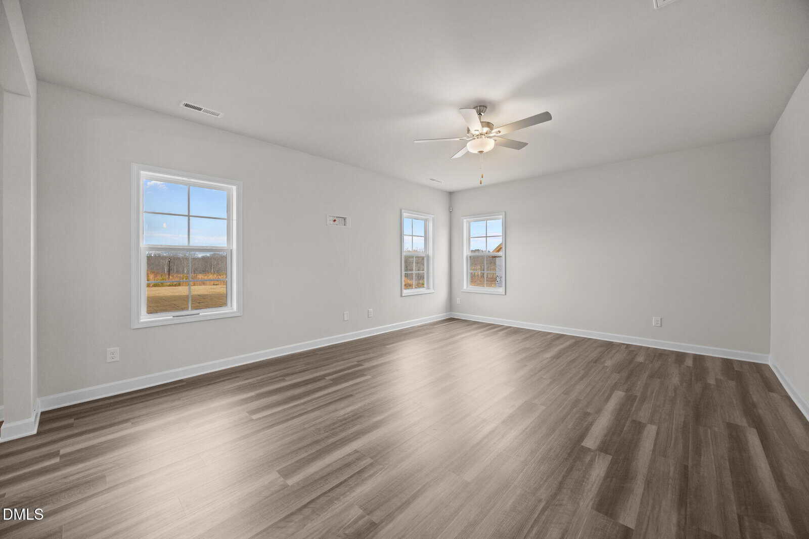 Spacious, empty room with light-colored walls, wood-style flooring, two large windows, and a ceiling fan with lights. Natural light streams in, highlighting the clean, modern interior.