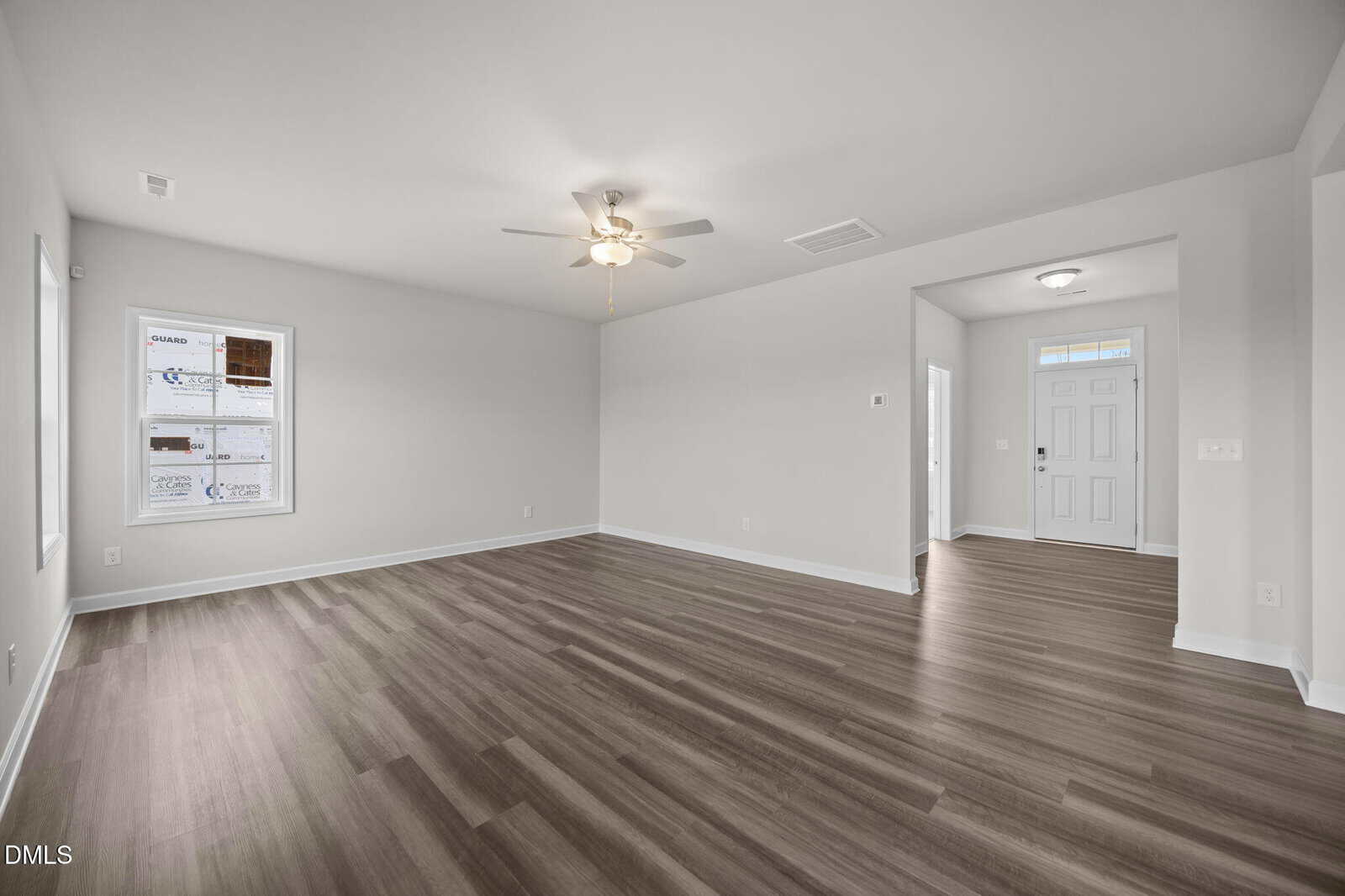 A spacious, empty living room with light gray walls, wood-look flooring, a ceiling fan with light, a window on the left, and an open entryway leading to a white front door.