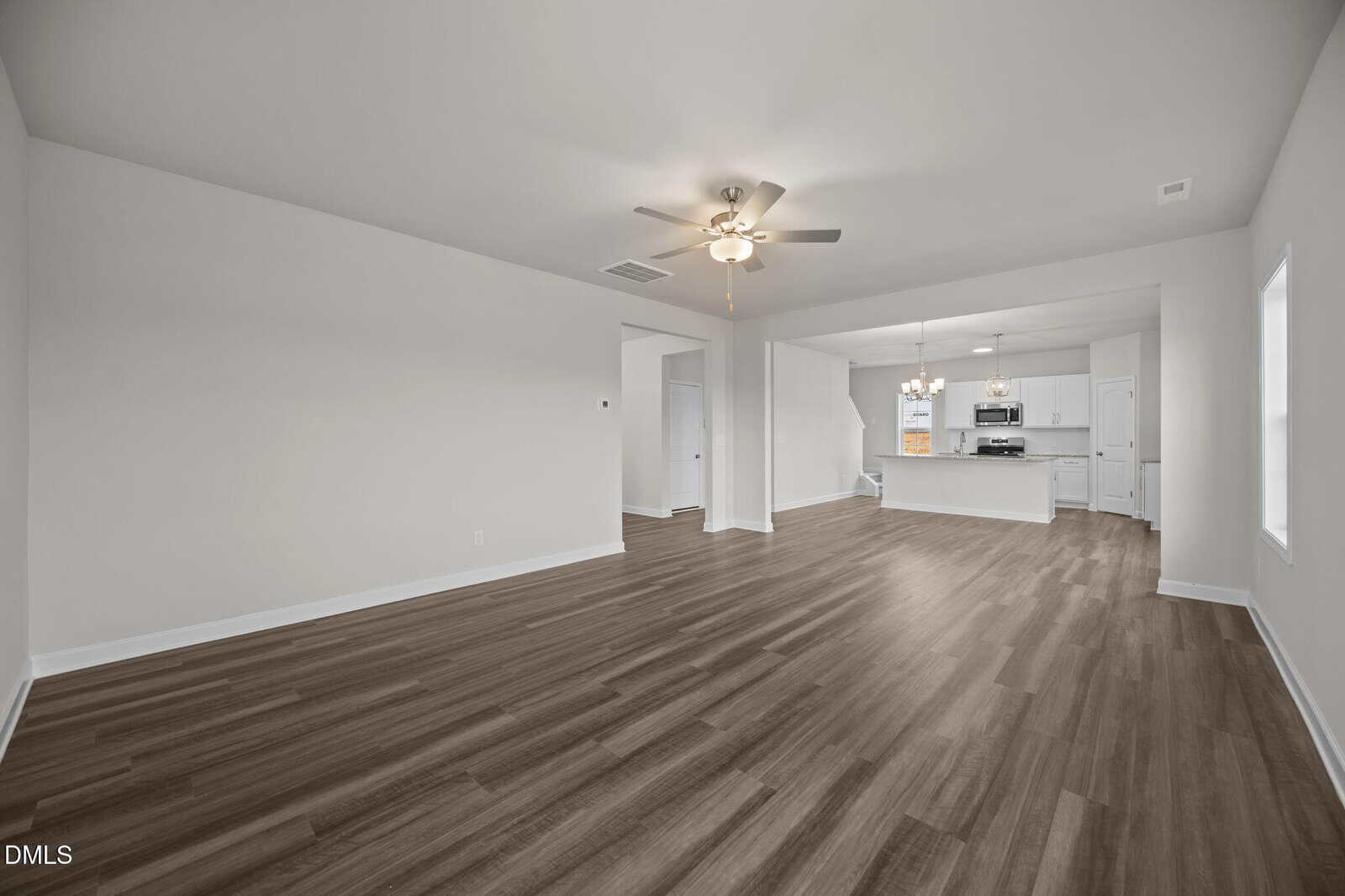 Spacious open-concept living area with wood-look flooring, neutral walls, ceiling fan, and view into a modern kitchen with white cabinets and stainless steel appliances.