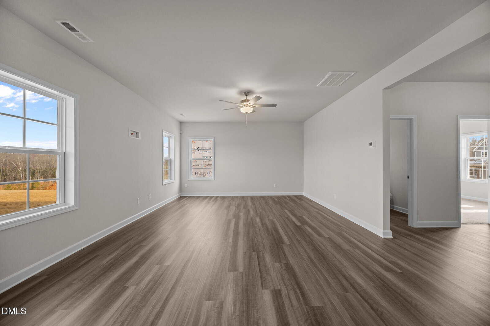 A spacious, empty living room with light gray walls, wood-look flooring, large windows, a ceiling fan, and a doorway leading to a bright adjacent room. Natural light fills the space from multiple windows.