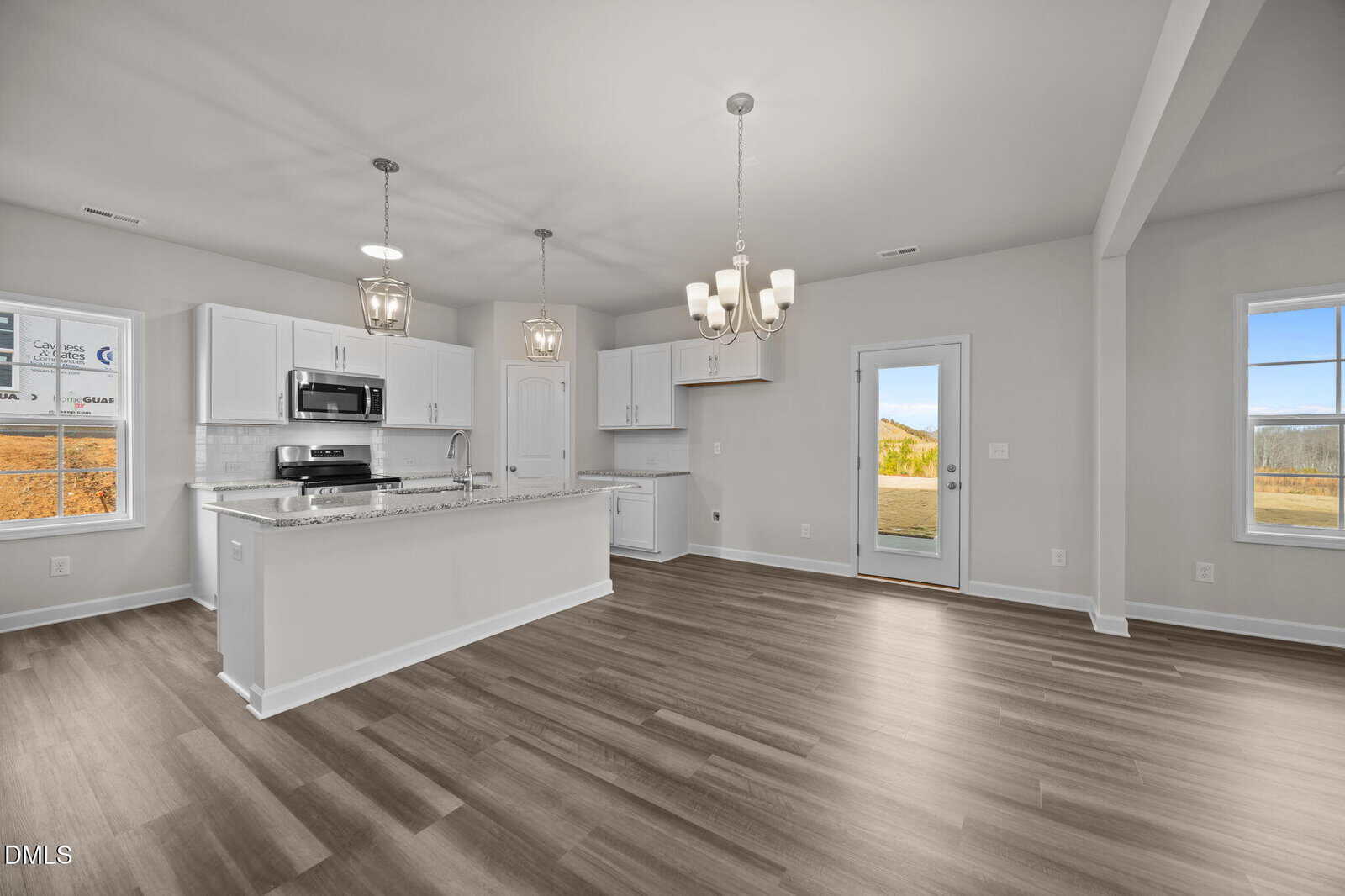 Modern kitchen with white cabinets, stainless steel appliances, an island with overhead pendant lights, and a dining area with a chandelier. Large windows and a glass door offer views of greenery and mountains outside.