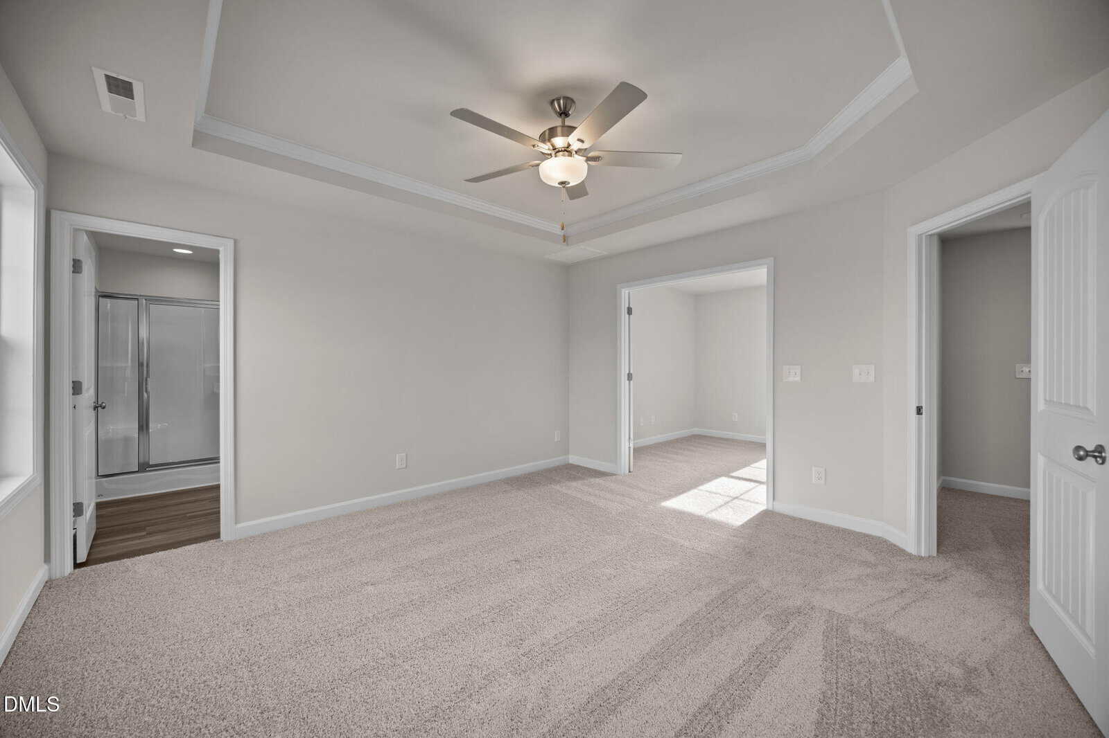 Spacious empty bedroom with beige carpet, tray ceiling with ceiling fan, two open doorways, and a window letting in natural light. A bathroom with a glass shower is visible through one doorway.