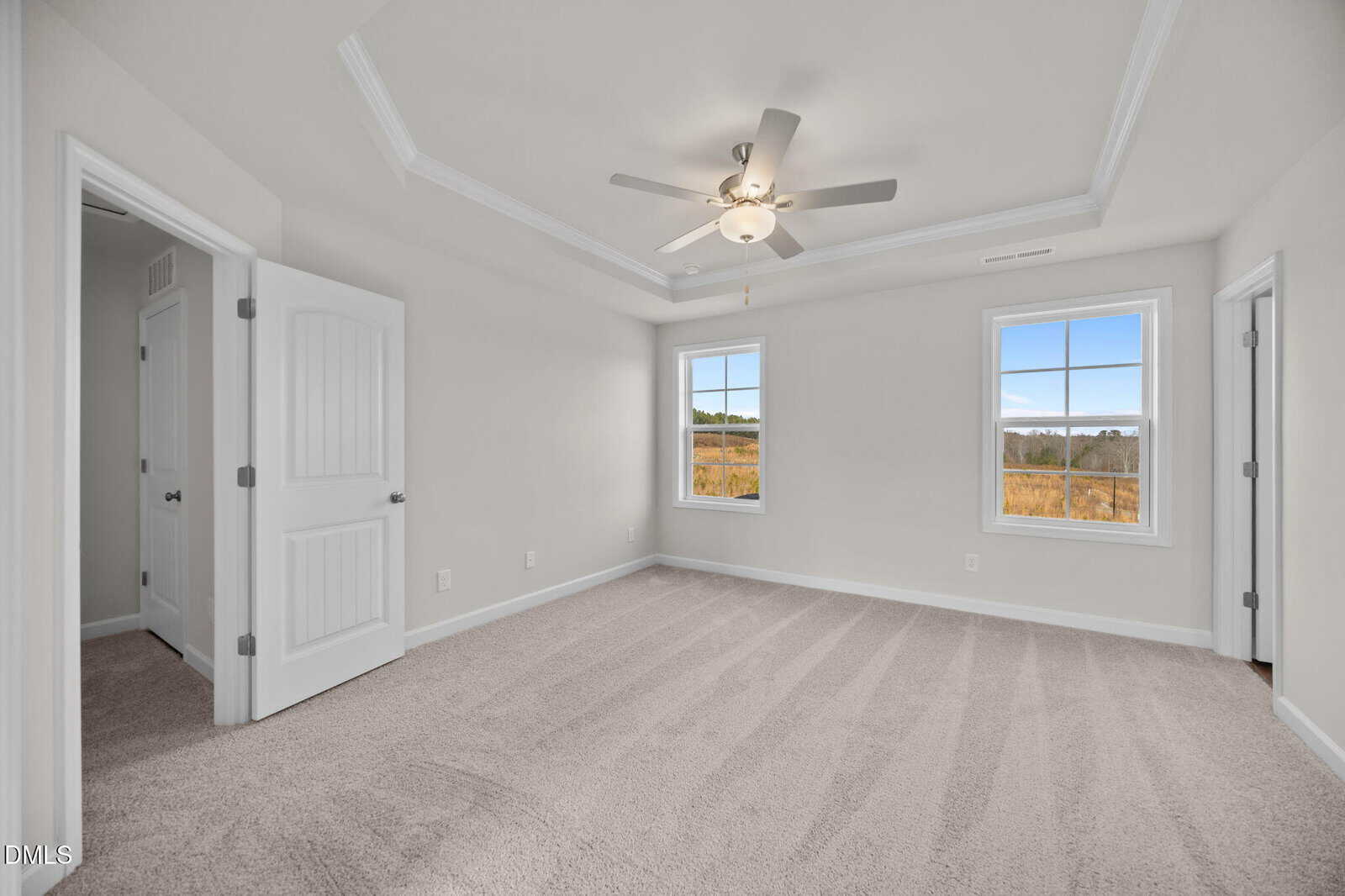 Bright, empty bedroom with beige carpet, white walls, ceiling fan, tray ceiling, two windows with outdoor view, and two open doors.