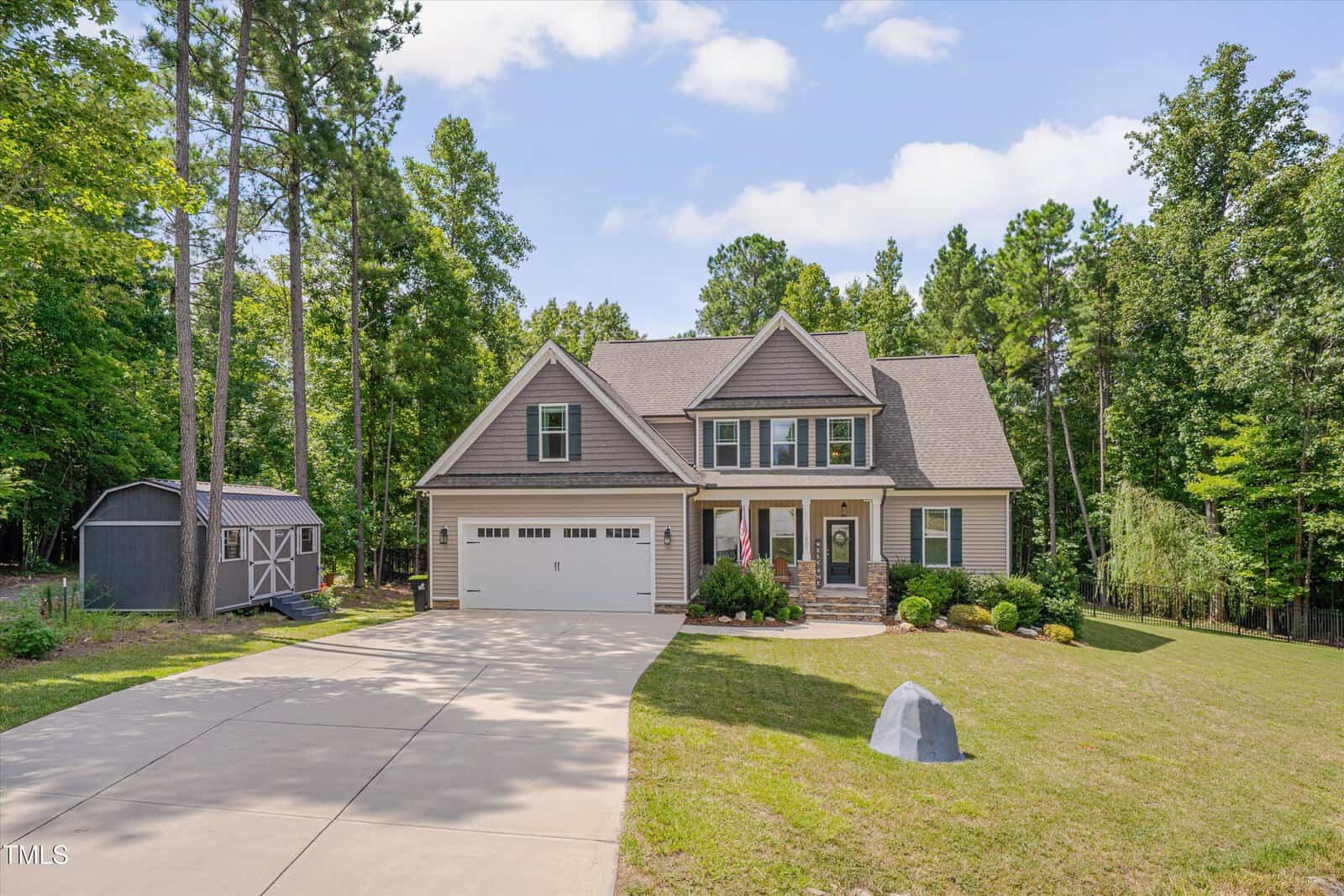 A two-story suburban house with gray siding, a gable roof, and an attached double garage is shown with a neatly landscaped front yard, a concrete driveway, and a gray storage shed on the left. Tall trees surround the property.