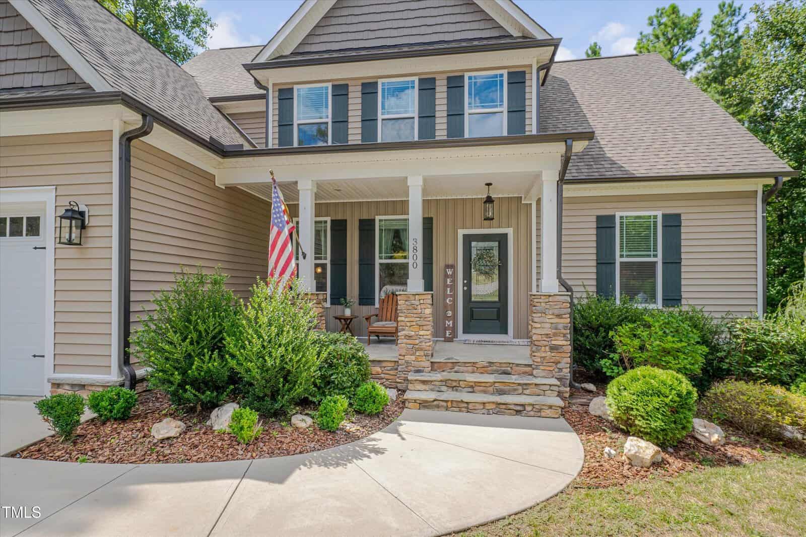 A two-story house with tan siding, stone porch columns, and white trim. The front porch has a welcome sign, two chairs, and an American flag. The landscaped yard includes bushes and a curved walkway.