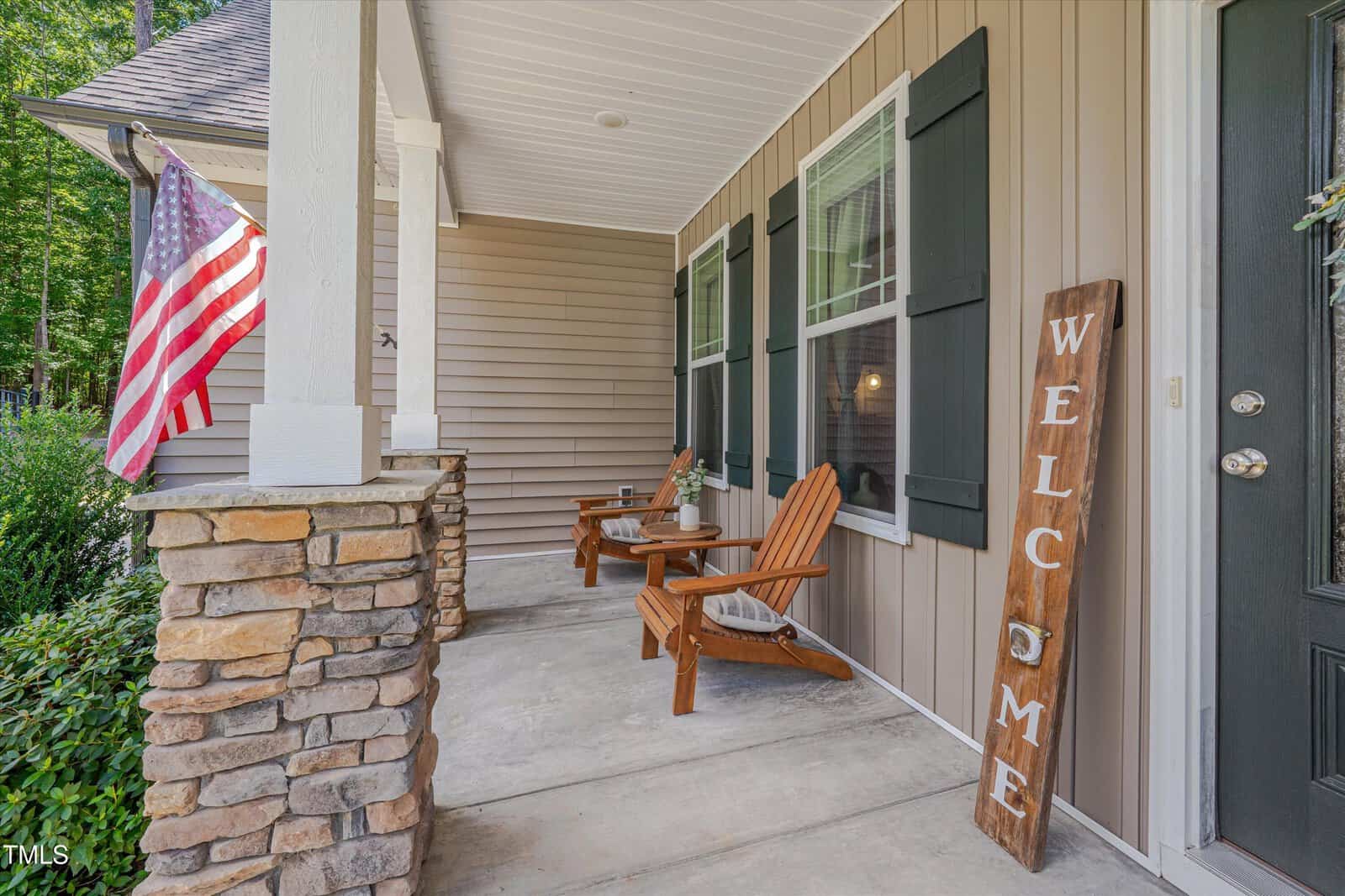 A front porch with stone columns, two wooden chairs, a small table, a large "Welcome" sign, and an American flag. The house has tan siding, black shutters, and greenery visible in the background.