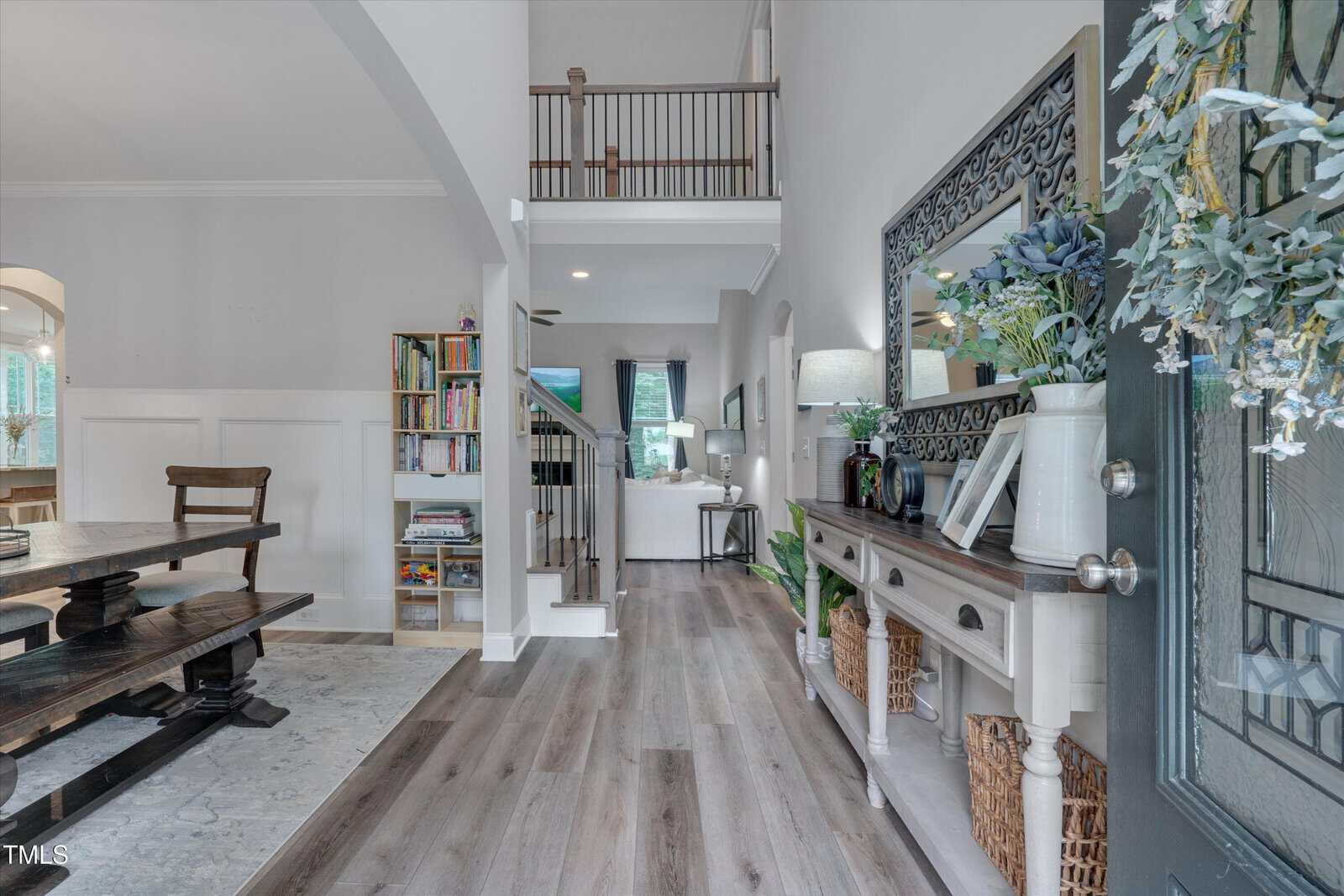 A view of a home entryway with light wood floors, a console table with decor and baskets, a staircase leading upstairs, bookshelves, a dining area to the left, and a living room visible in the background.