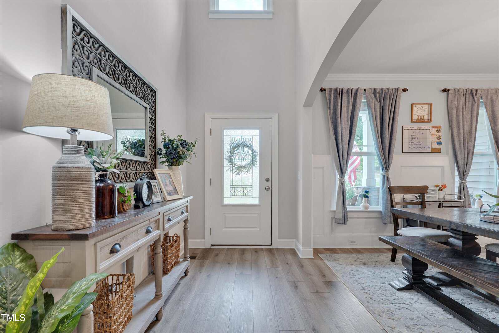 A bright entryway with a white front door, a sideboard table holding a lamp, baskets, and framed photos. To the right, a dining area with a wooden table and chairs is visible, featuring blue curtains and wall decor.