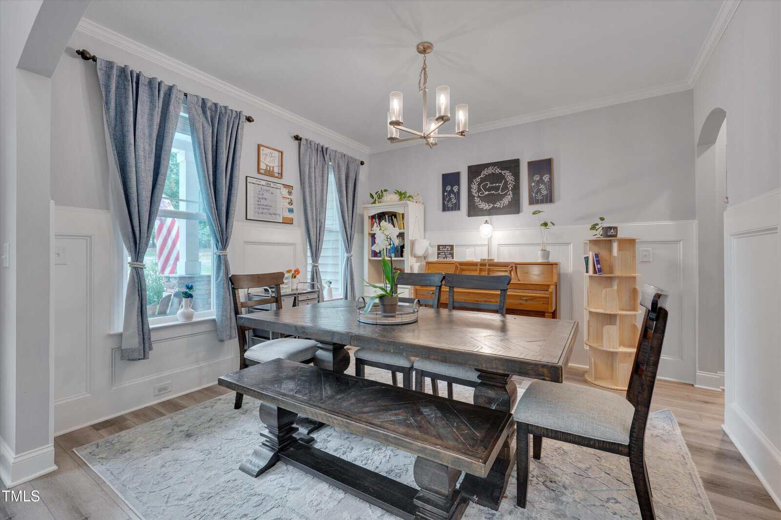 A dining room with a dark wooden table and bench, four chairs, light gray walls, and hardwood floors. Large windows with blue curtains, a chandelier, wall art, a piano, bookshelf, and houseplants are visible.
