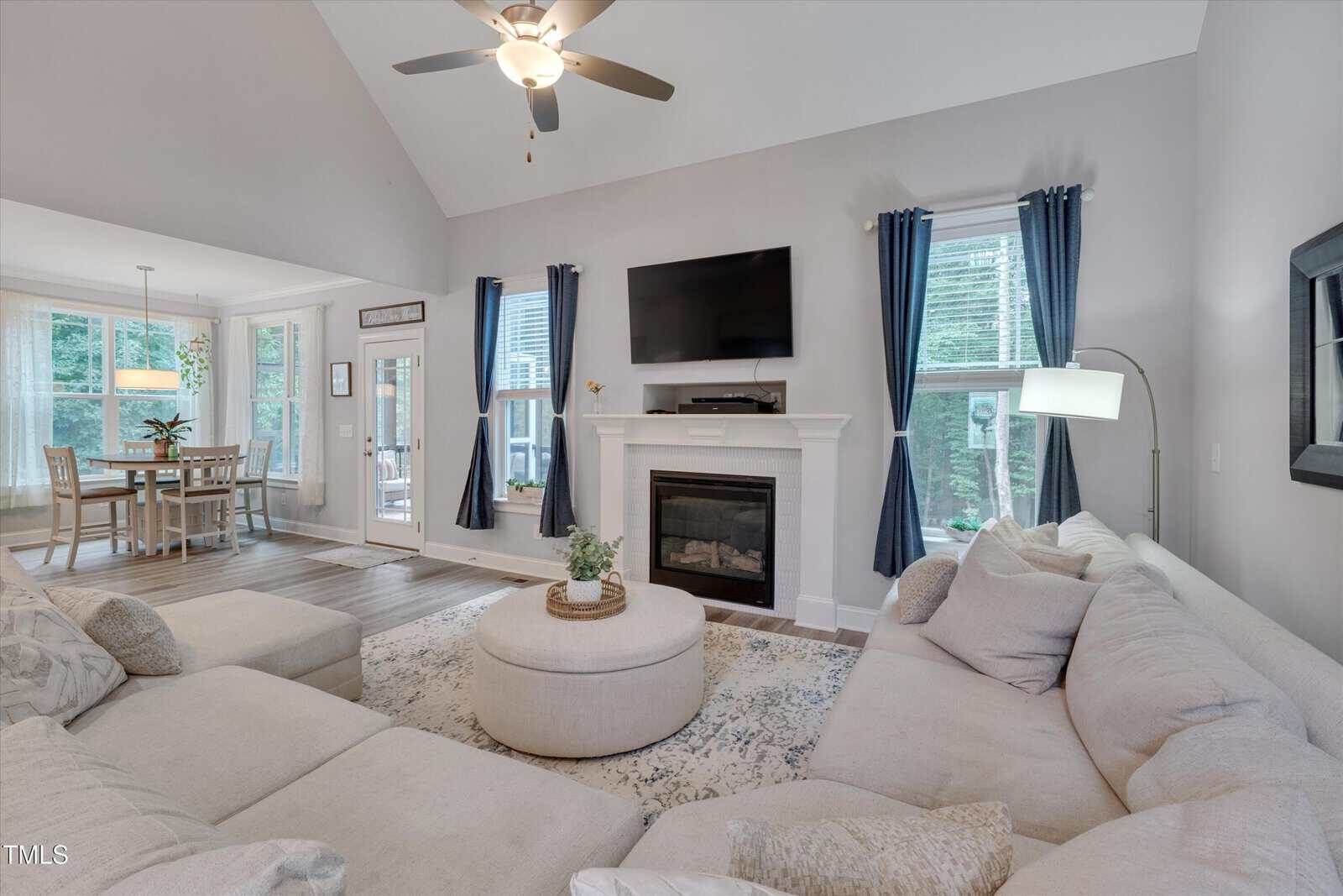 A spacious living room with a beige sectional sofa, round ottoman, and patterned rug. There is a fireplace with a TV mounted above it, large windows with blue curtains, and a ceiling fan. A dining area is visible in the background.