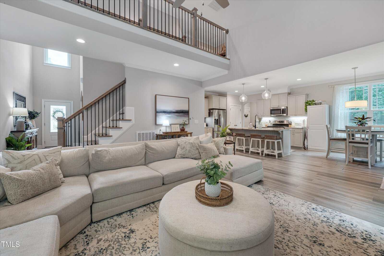 Spacious open-concept living area with a large beige sectional sofa, round ottoman, and kitchen with white cabinets and an island. Wooden stairs and a balcony railing are visible in the background. Natural light fills the room.