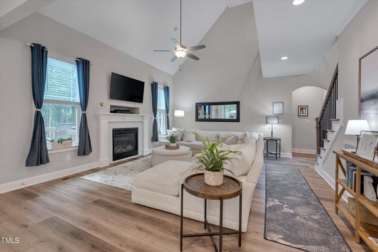 A modern living room with light wood floors, a white sectional sofa, a fireplace with a wall-mounted TV above it, large windows with blue curtains, and a small round side table with a potted plant.