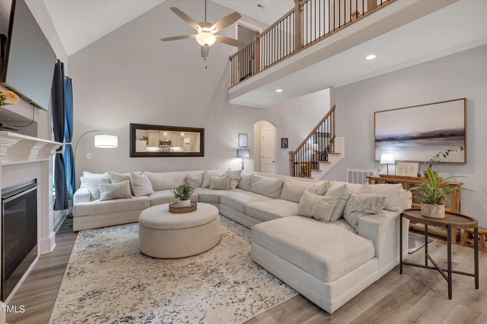 Spacious living room with a large L-shaped beige sectional, round ottoman, patterned rug, fireplace, plants, and stairs leading to an open loft above. Neutral tones and modern decor create a cozy, inviting space.