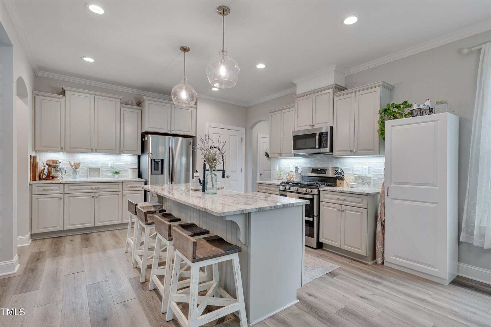 Modern kitchen with white cabinets, stainless steel appliances, marble island with four wooden stools, pendant lights, light wood flooring, and decorative plants. The space appears bright, clean, and inviting.