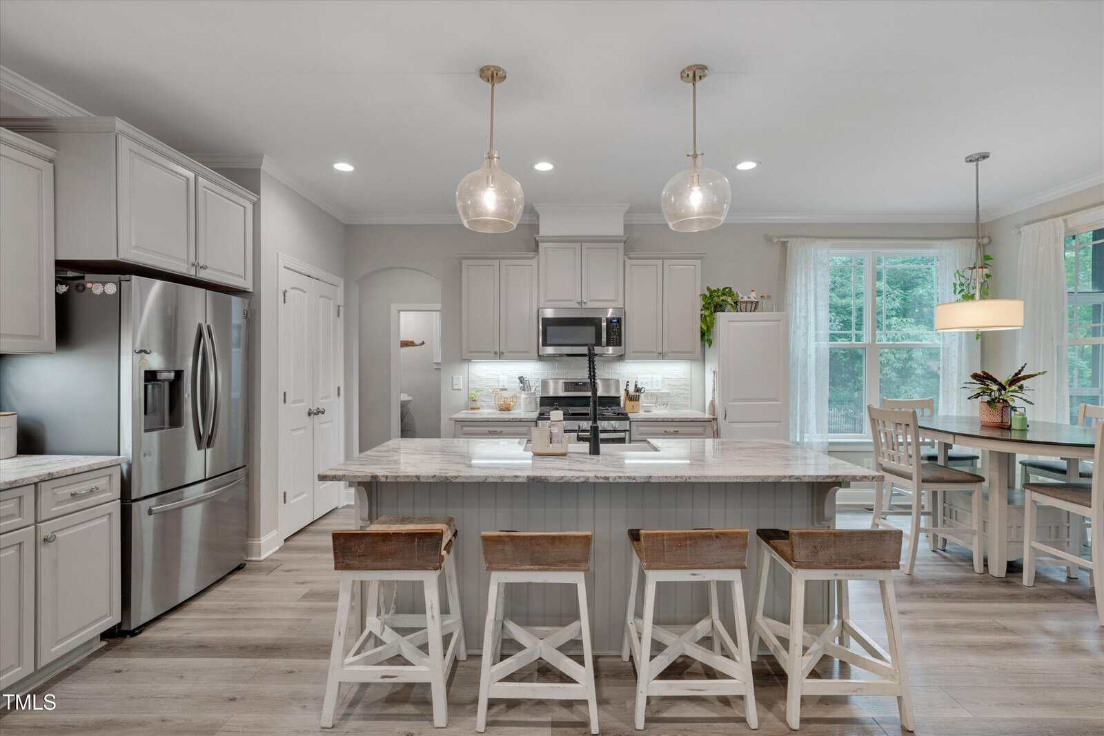 Modern kitchen with a large island, three wooden stools, stainless steel appliances, pendant lights, and a dining area by large windows with sheer curtains and a small plant on the table.