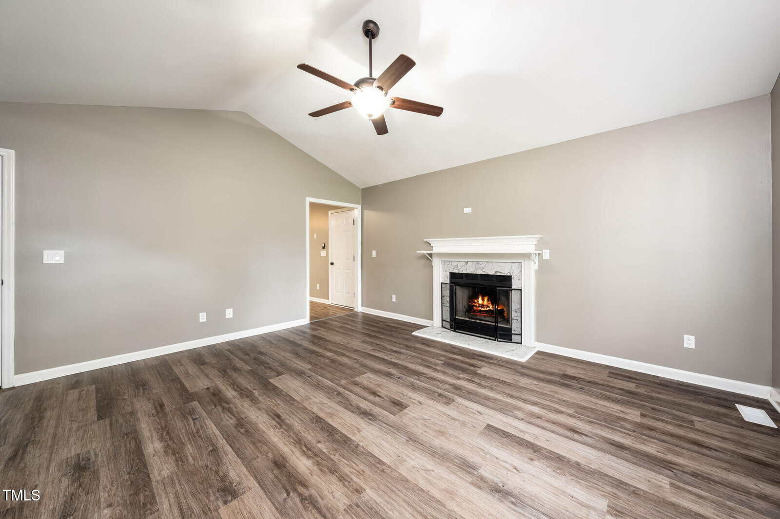 A spacious living room with light gray walls, wood-look flooring, a ceiling fan, and a white fireplace with a lit fire. The room is empty and has an open doorway leading to another area.