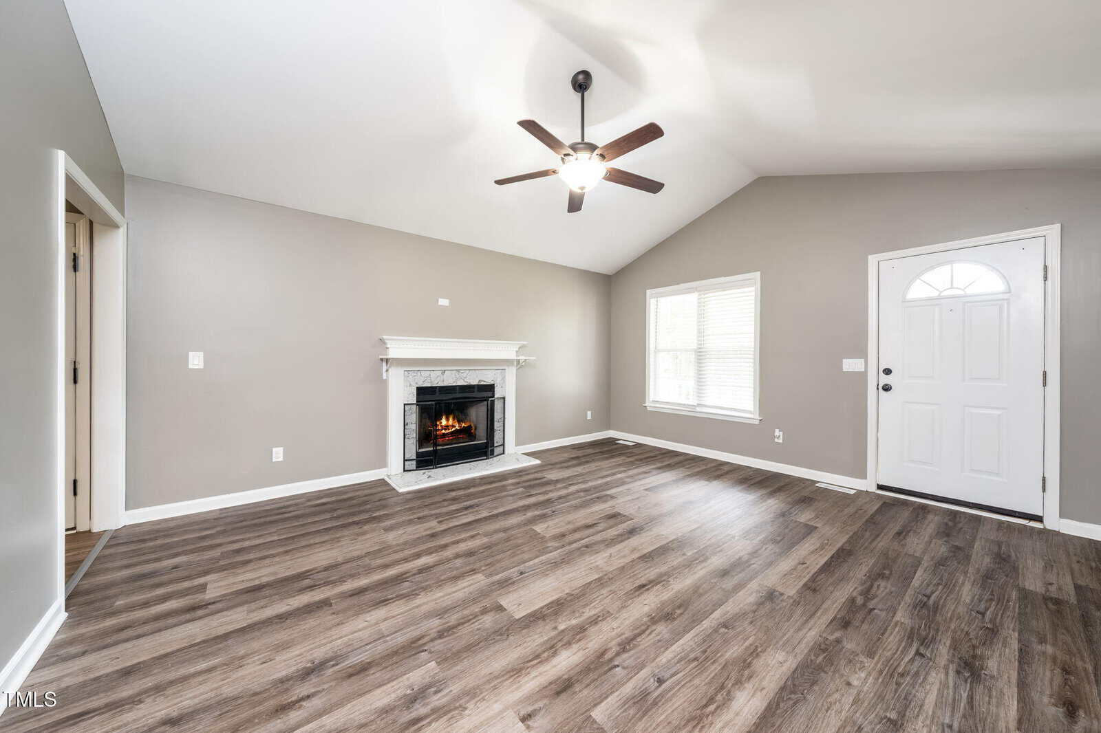 A spacious living room with light gray walls, wood-style flooring, a white front door, a large window, a ceiling fan with a light, and a white fireplace with a mantel. The room is empty and well-lit.