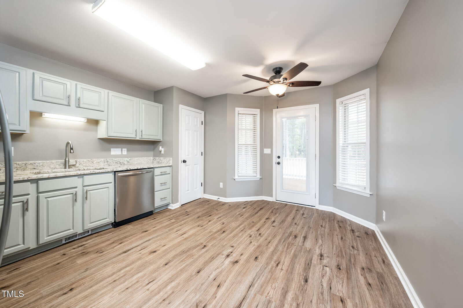 Bright kitchen with light gray cabinets, granite countertops, stainless steel dishwasher, wood-look flooring, a ceiling fan, and double doors leading outside. Two windows provide additional natural light.