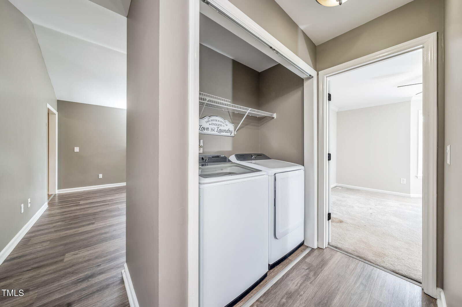 Open closet with a washer and dryer in a hallway, adjacent to rooms with wood and carpeted floors; walls are painted light taupe, and a sign on the shelf reads “Love & Laundry.”.