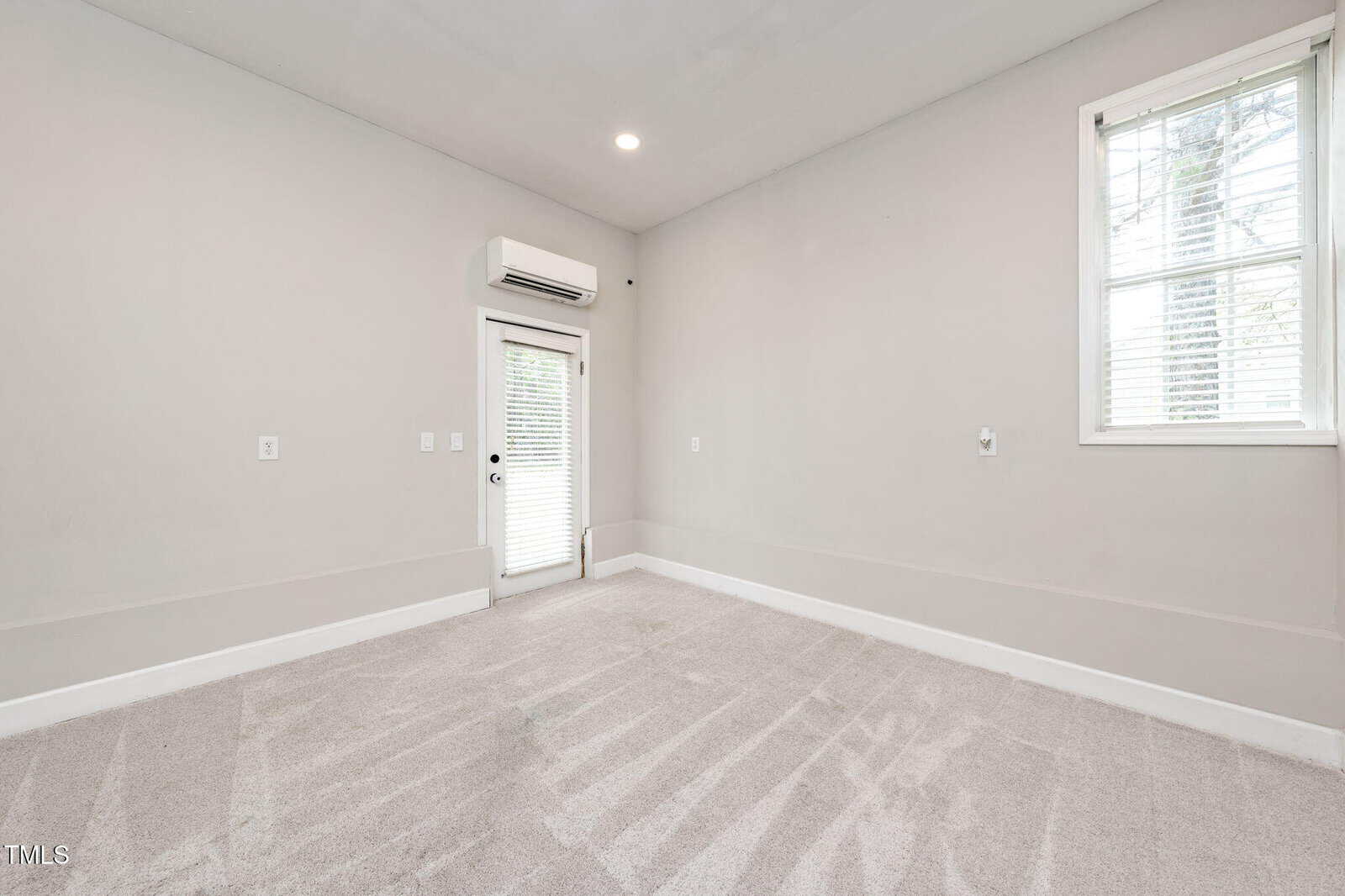 Empty, light gray carpeted room with white walls, a window with blinds, a glass door, and a wall-mounted air conditioning unit above the door. Sunlight is streaming in through the window.