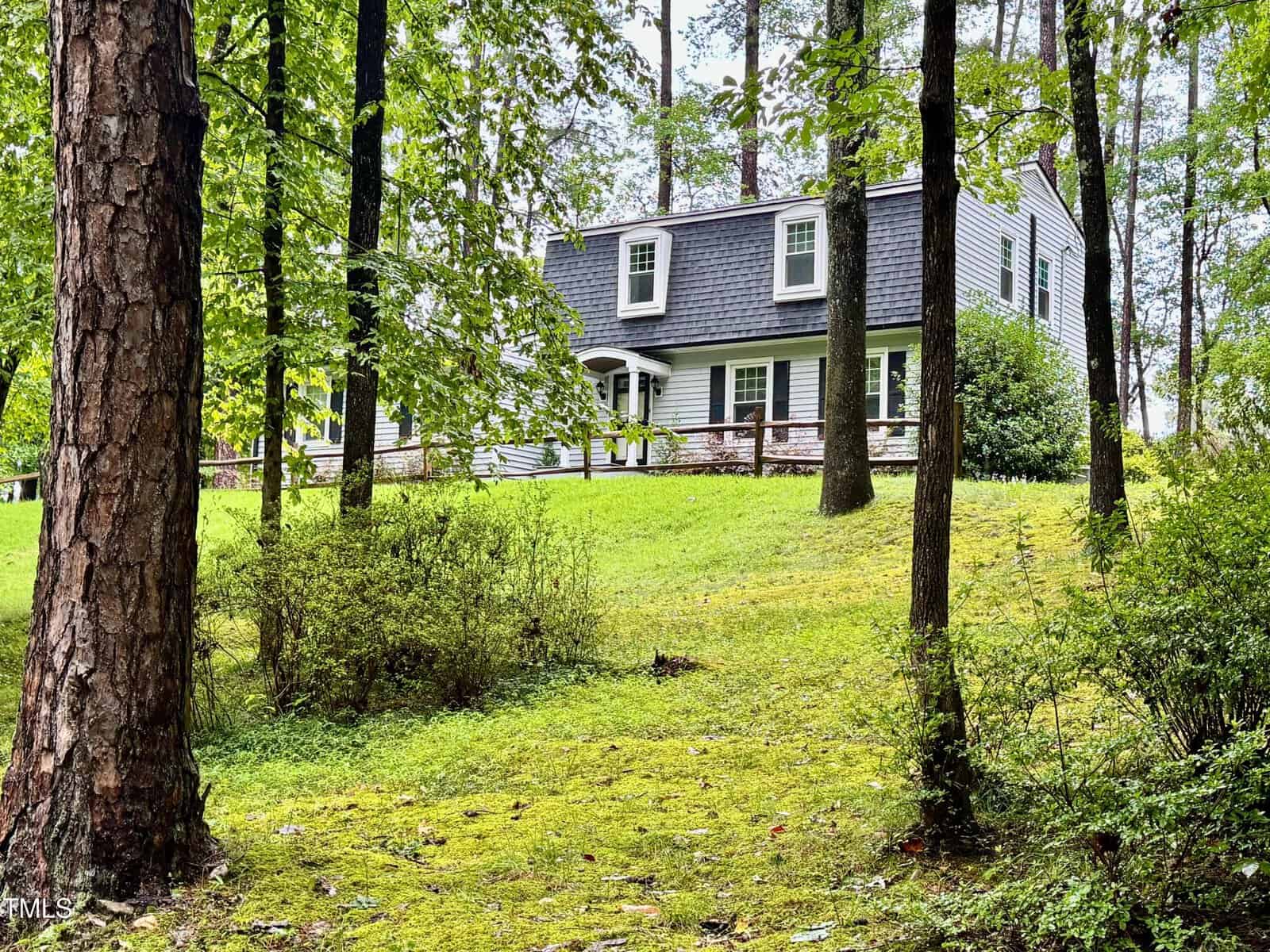 A two-story house with a dark shingles roof, white siding, and dormer windows sits on a grassy, wooded lot with several tall trees and a wooden handrail along the front porch.