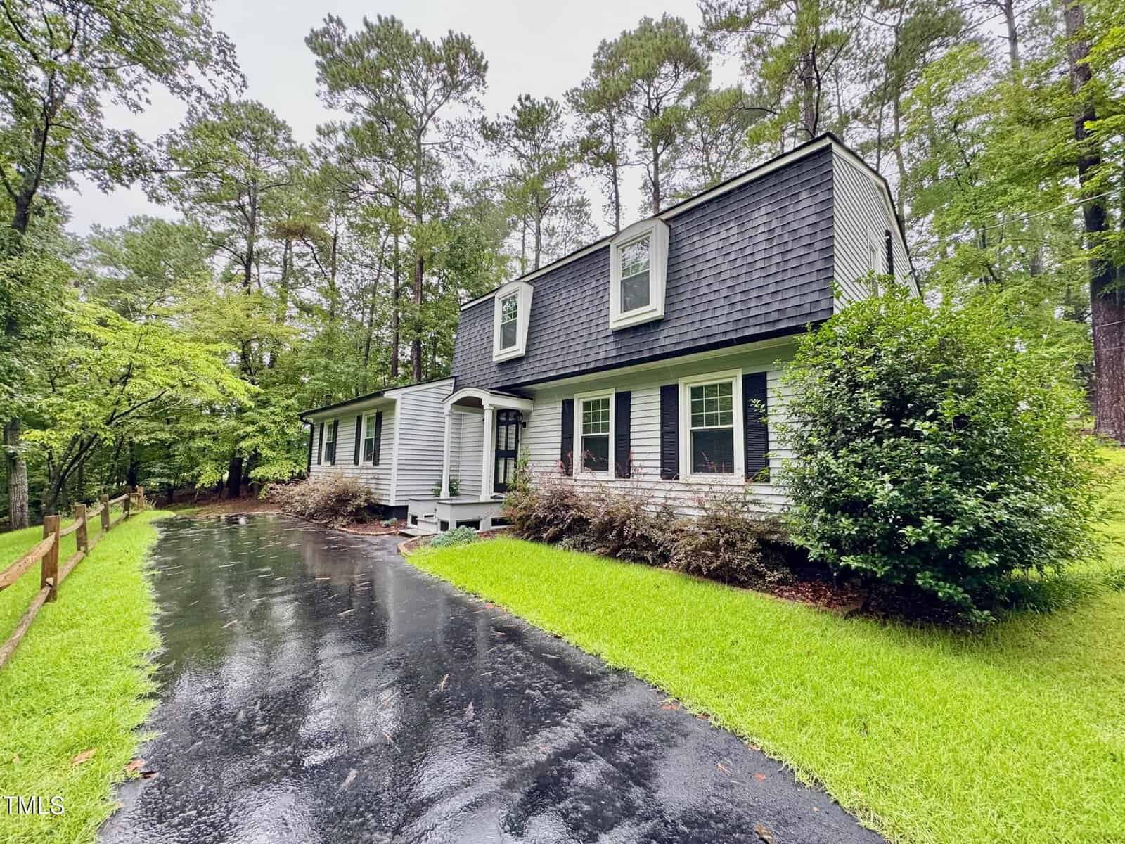 A two-story house with a dark gray gambrel roof and white siding sits beside a wet asphalt driveway, surrounded by green grass, bushes, and tall pine trees on a cloudy day.
