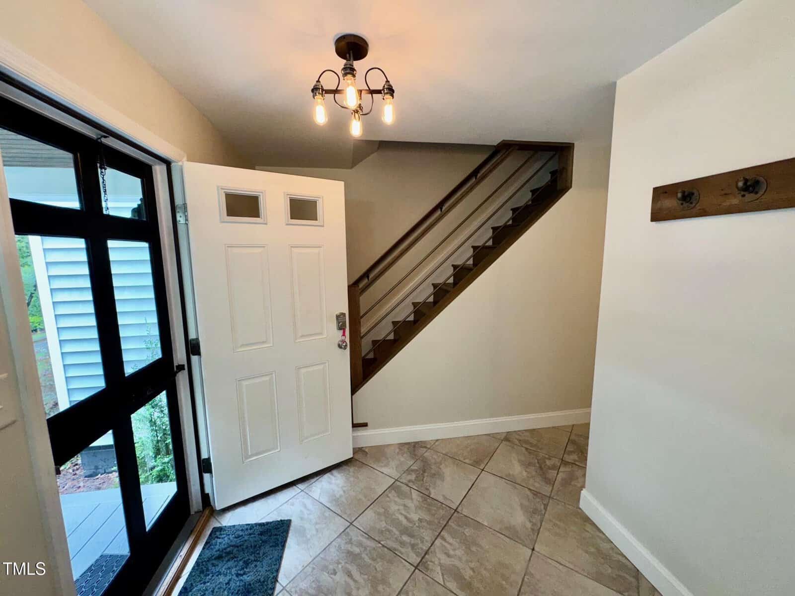 A bright entryway with light tile flooring, an open white front door, a staircase with dark wooden trim, a wall-mounted coat rack, and a ceiling fixture with three exposed bulbs.