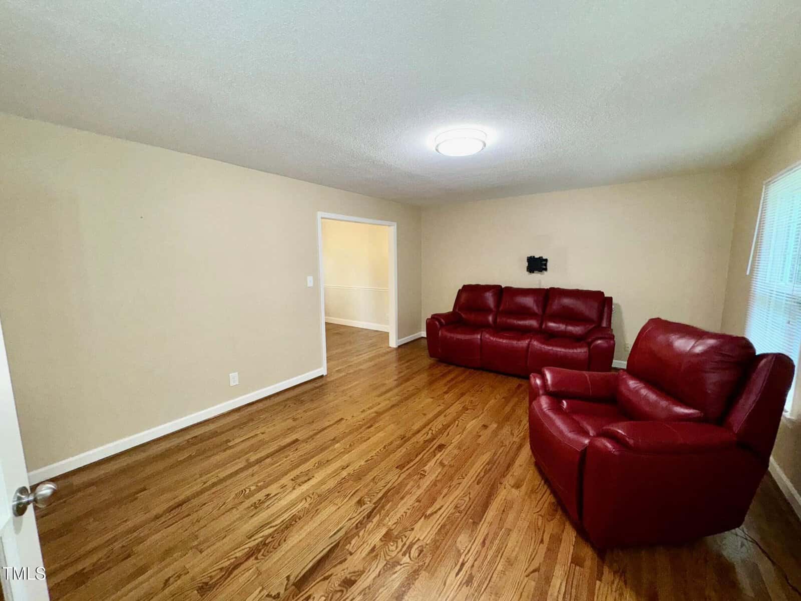 A living room with hardwood floors, beige walls, a ceiling light, a red leather sofa, and a matching red armchair. A doorway leads to another room, and two windows let in natural light.