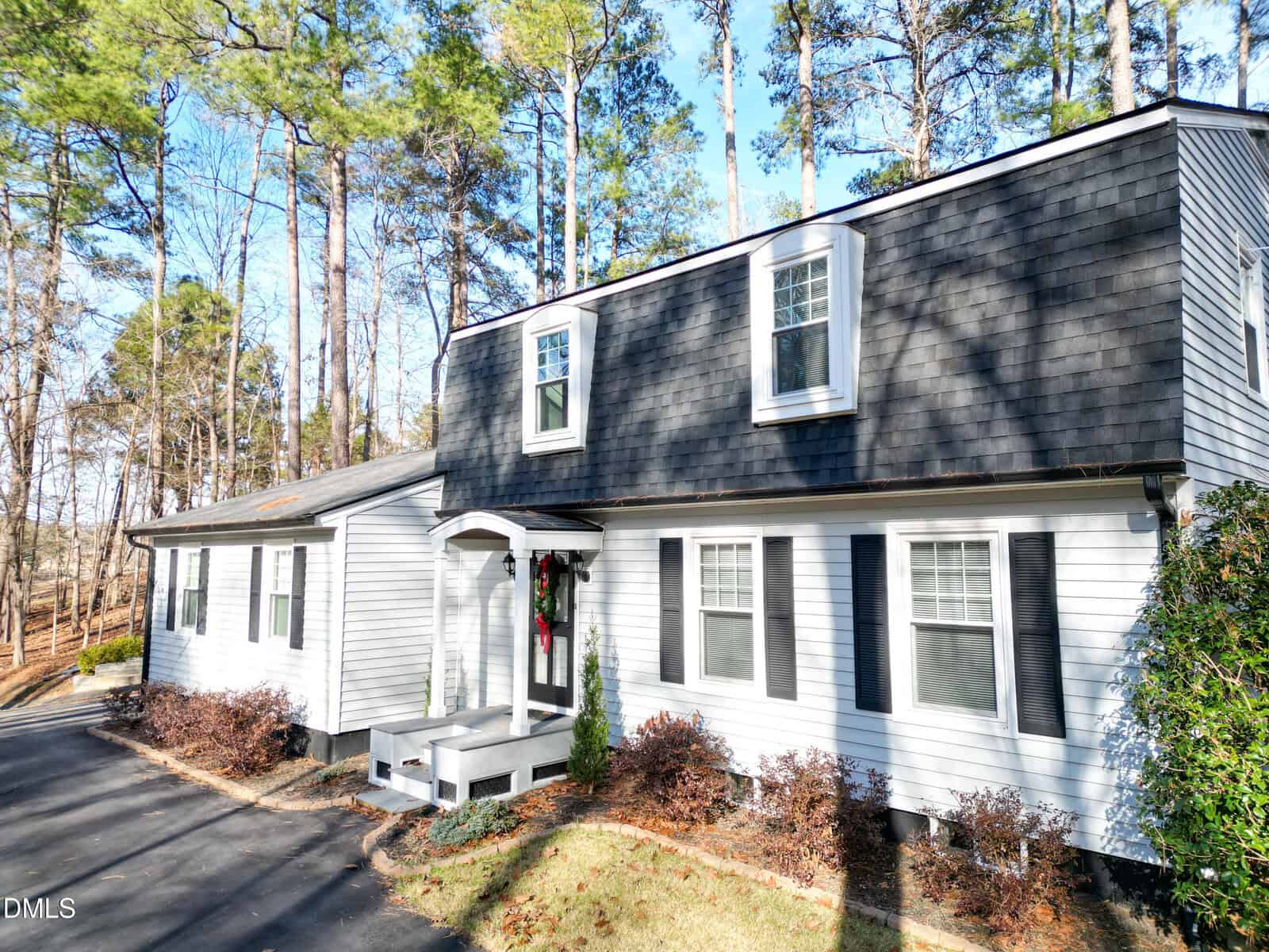 A two-story house with gray siding and dark gray shingles, surrounded by trees. The home has white trim, black shutters, and a small front porch with a wreath on the door. A driveway runs alongside the house.