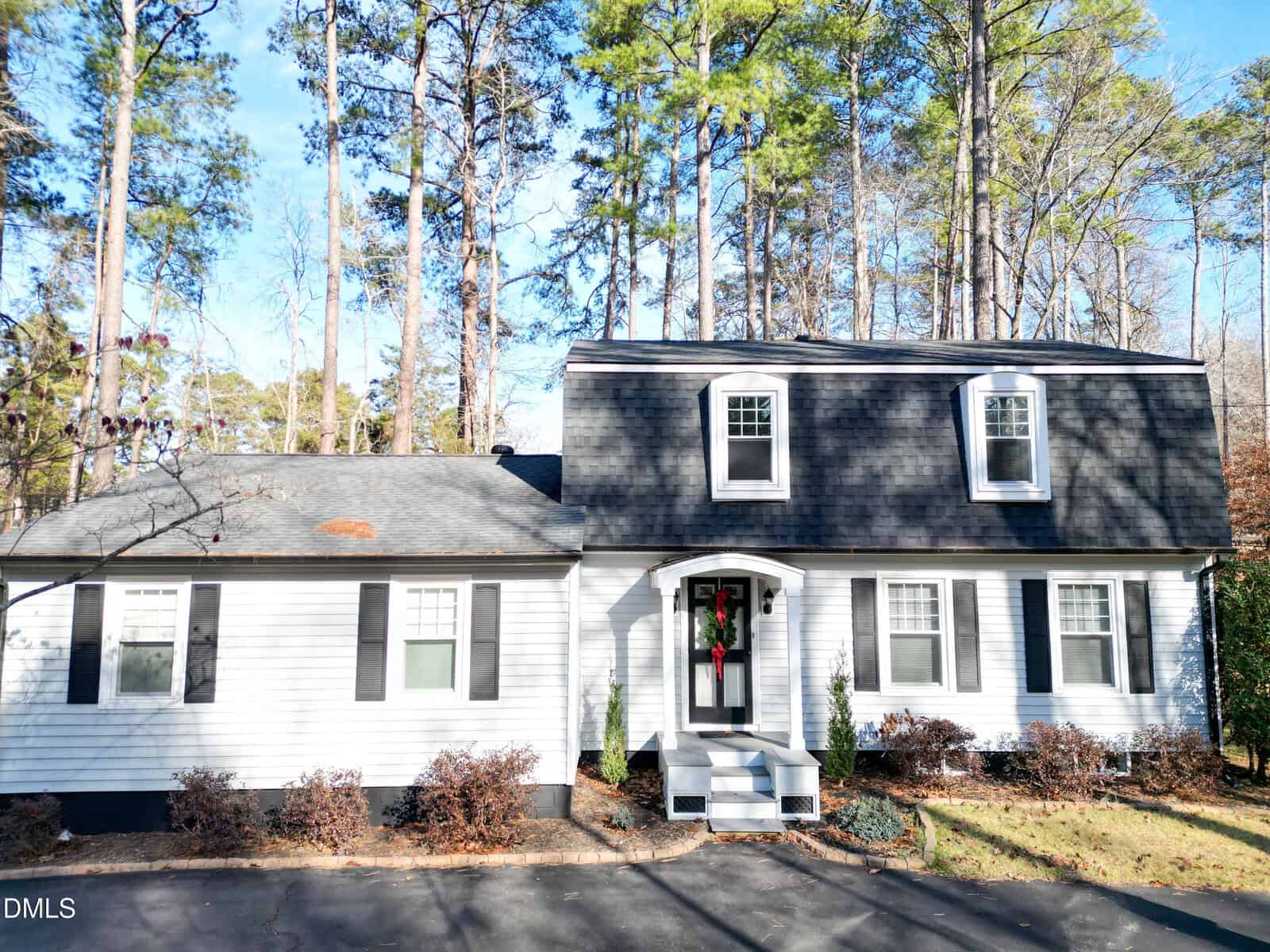 A two-story white house with black shutters and a dark roof, featuring three dormer windows on the upper floor. There is a small front porch with a wreath on the door, surrounded by trees and shrubs.
