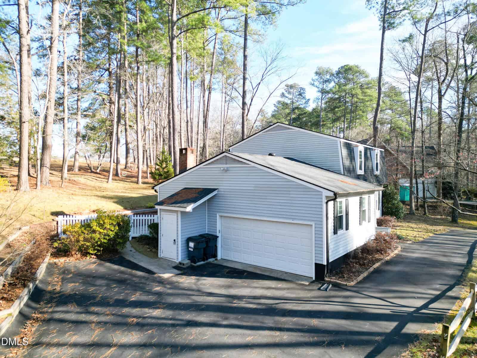 A two-story white house with a gray roof, attached two-car garage, and driveway. The home is surrounded by trees and has a white fence and shrubs along one side. The sky is clear and sunny.